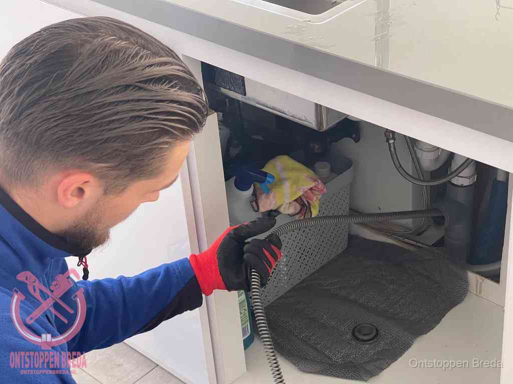 Een monteur voert een flexibele ontstoppingsveer in onder het keukenkastje, langs de sifon, om de gootsteen te ontstoppen in Breda Een monteur voert een flexibele ontstoppingsveer in onder het keukenkastje, langs de sifon, om de gootsteen te ontstoppen in Breda