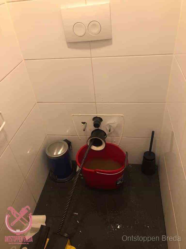 Een ontstopte wand-wc met verwijderde toiletpot waar een ontstoppingsveer in de wanduitgang verdwijnt, het achterhalen waar de verstopping zit in Breda