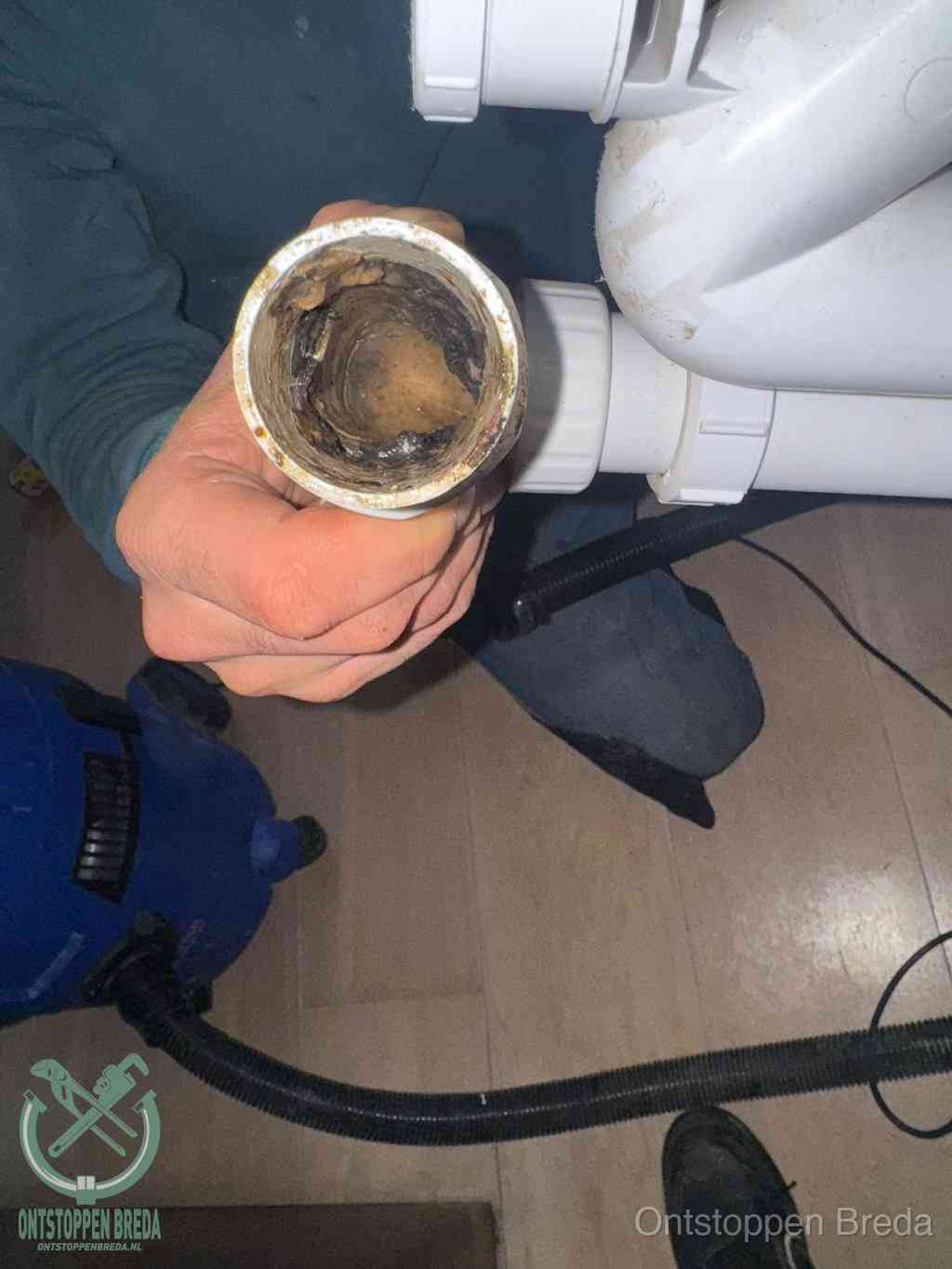 Een hand houdt een losgekoppeld PVC-afvoerbuisstuk vast dat van binnen volledig is dichtgeslibd met vet, vuil en aanslag, de meest voorkomende oorzaak van een verstopte afvoer in Breda