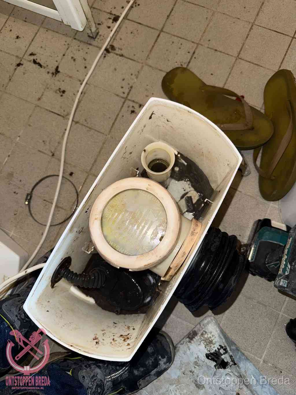 Een gedemonteerde toiletpot op de vloer met ontstoppingsveer en gereedschap ernaast, het verwijderen van een vastzittend voorwerp uit de wc in Breda